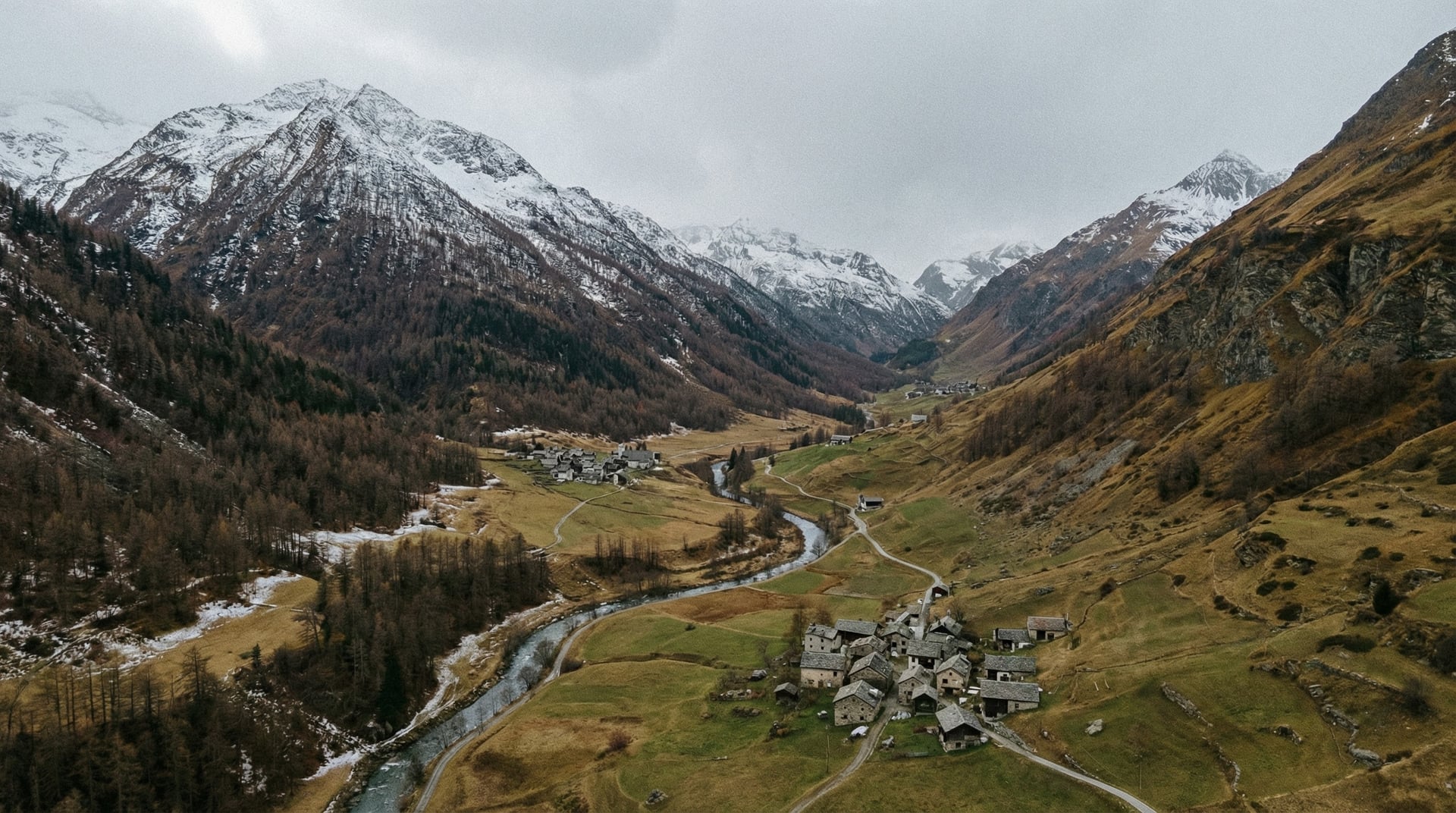 Luftaufnahme eines Alpentals mit gewundenem Fluss, Steindorf und schneebedeckten Gipfeln unter grauem Himmel