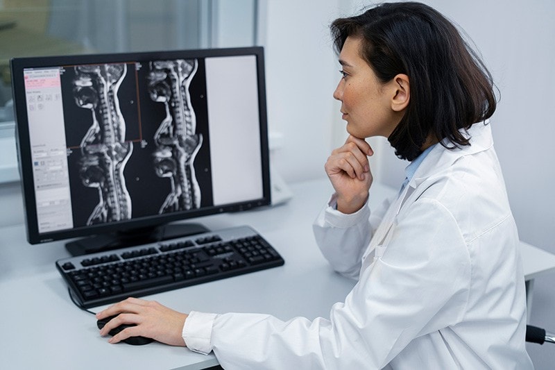 Médecin en blouse blanche examinant des images IRM de la colonne vertébrale sur un écran de bureau en clinique